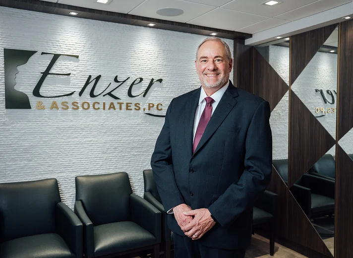 A professional headshot of Dr. Enzer, a man in a dark suit and tie, smiling and standing in the waiting area of his office, Enzer & Associates, PC.
