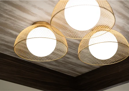 A close-up view of three modern ceiling light fixtures with round, glowing white bulbs housed inside dome-shaped gold wire mesh cages, set against a wooden ceiling.