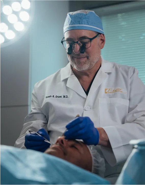 Dr. Yoash R. Enzer, M.D., wearing a surgical cap, loupes, and a white lab coat, is leaning over a patient covered in surgical drapes, preparing for or performing a procedure with instruments in his gloved hands.