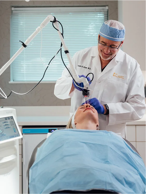 Dr. Enzer, wearing a surgical cap and lab coat, administers a laser treatment to a patient's nose area using a device with an articulating arm. The patient is lying down covered by a blue drape.