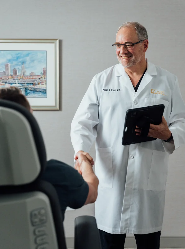 Dr. Yoash R. Enzer, M.D., wearing a white lab coat and holding a tablet, smiles while shaking hands with a patient seated in an examination chair. A framed watercolor painting hangs on the wall behind him.