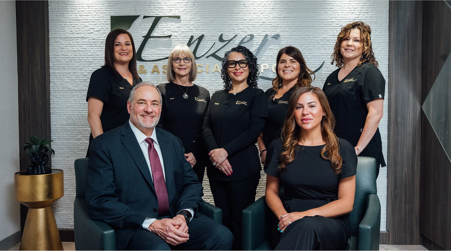 A group portrait of Dr. Enzer and six female team members. Dr. Enzer is seated in the front left in a suit, and the team members are standing or seated around him, all wearing black tops, in the office waiting area with the Enzer & Associates, PC logo visible behind them.
