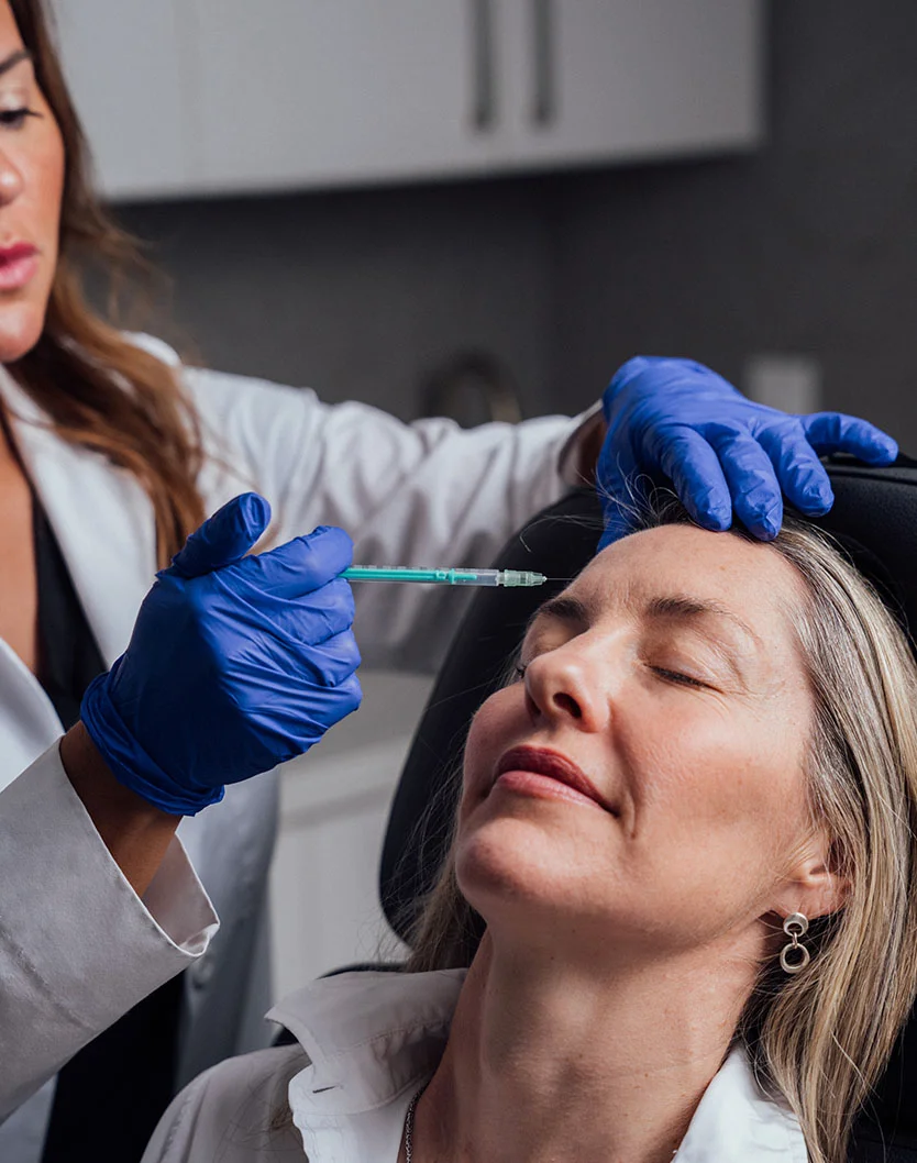 A close up of a female medical professional in a white lab coat and blue gloves is administering an injectable treatment, holding a syringe near the forehead of a relaxed female patient who is seated in a treatment chair. - Botox Dysport Daxxify in Providence, RI