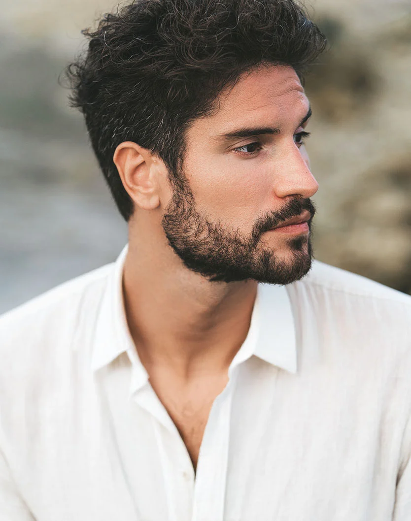 A close-up profile portrait of a man with dark, curly hair and a beard, wearing an unbuttoned white collared shirt, looking off to the right. - Earlobe Repair in Providence, RI