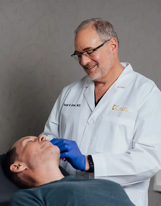 Dr. Enzer, wearing a white lab coat and glasses, smiles while examining the neck and jawline of a male patient who is lying down in a treatment chair.