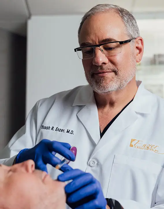 A doctor in a white lab coat and blue gloves, Dr. Yoashi Enzer, M.D., is administering an injection to a patient lying down.