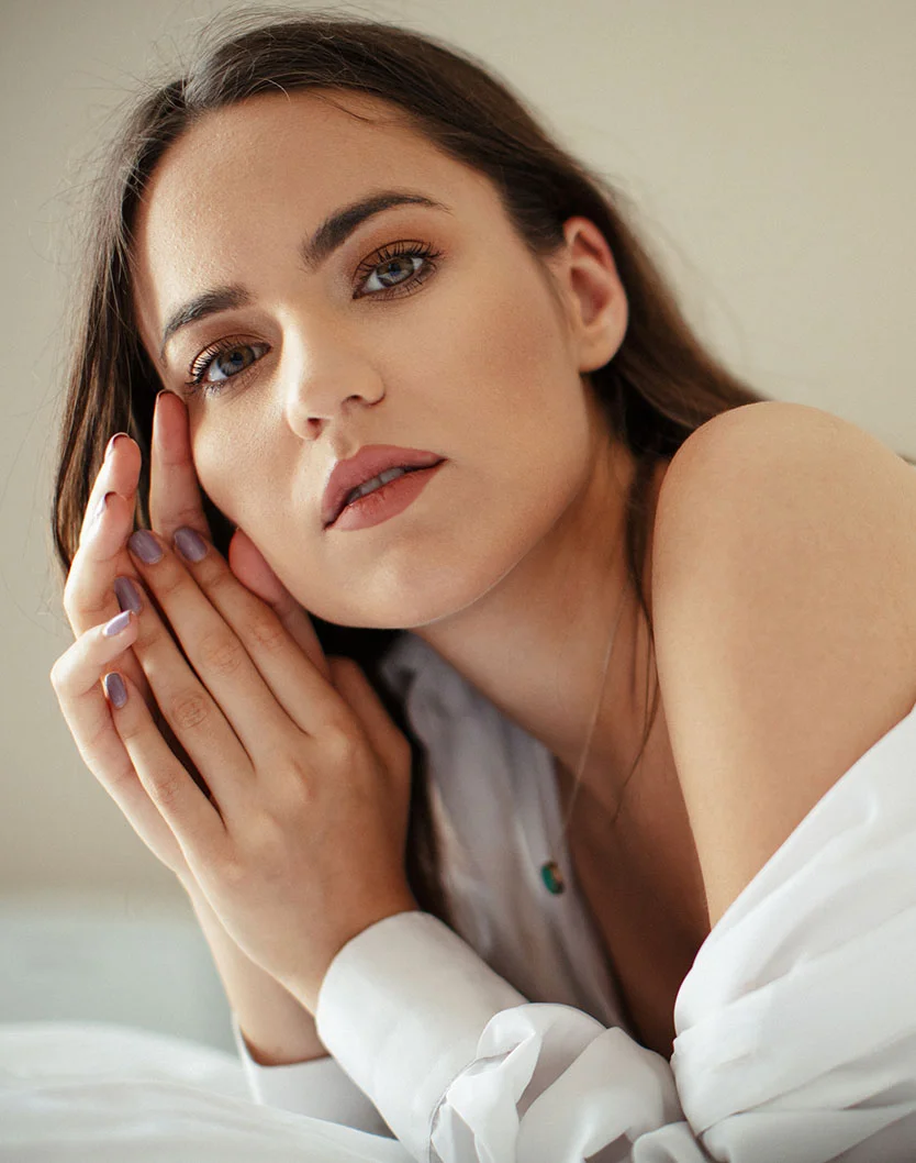 A close-up portrait of a young woman with long dark hair and striking eye makeup, resting her head on her hands and looking intently at the camera, while wearing a partially unbuttoned white shirt. - Eyelash Treatment in Providence, RI