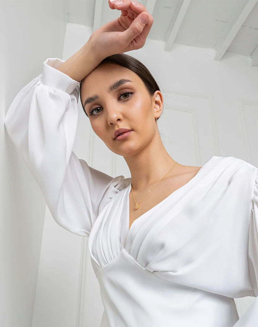A studio portrait of a woman with dark hair pulled back, wearing a white satin blouse with puffed sleeves and a delicate gold necklace, resting her arm against a white wall. - Eyeliner Micropigmentation in Providence, RI