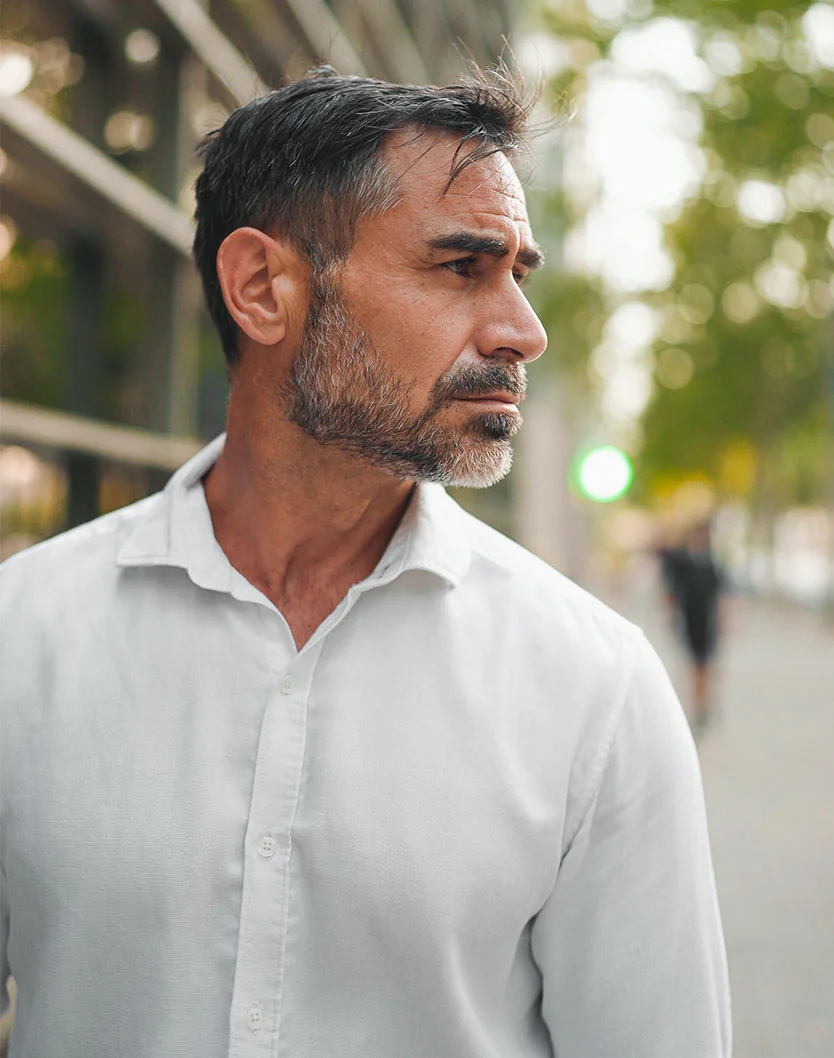 A mid-range close-up of a middle-aged man with short, dark graying hair and a neatly trimmed gray beard. He gazes off to his right with a neutral, thoughtful expression. He wears a pale off-white button-down shirt with the top buttons undone, slightly revealing his chest. The background is blurred, showing dark gray and black structures, possibly a modern building with glass walls, suggesting a business or urban setting. - Facial and Eylid Spasms in Providence, RI