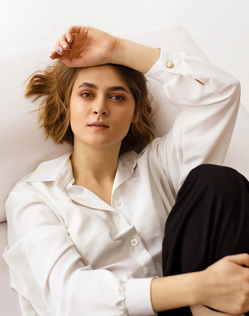 A portrait of a woman with short brown hair, wearing a white collared shirt and black pants, lying on a white surface with one arm resting on her forehead. - Facial Paralysis in Providence, RI
