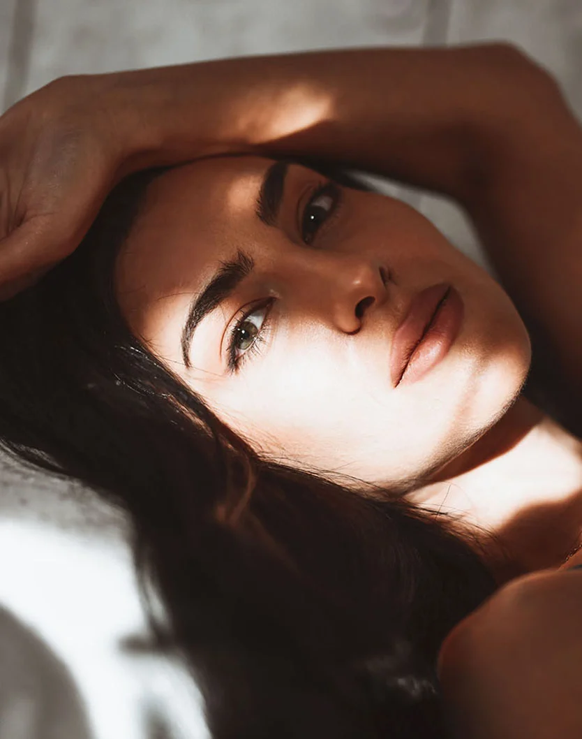 A woman with dark hair lies on a light beige surface with her arms extended overhead. Sunlight casts soft shadows across her face and body, creating a warm, intimate mood. She has a thoughtful expression and wears a light-colored necklace. The background is a light gray-beige wall. - Fat Grafting in Providence, RI