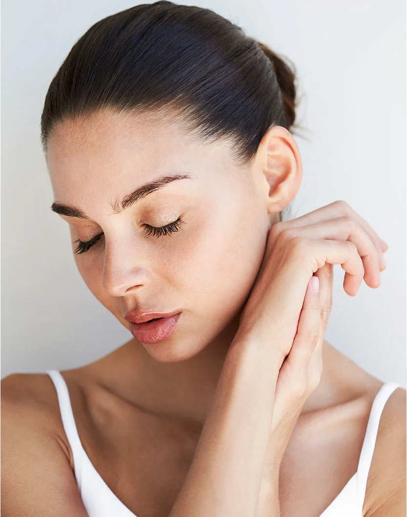 A close-up studio portrait of a woman with her eyes closed, dark hair pulled back, wearing a white tank top, gently touching her face with both hands. - Forehead Lifts in Providence, RI