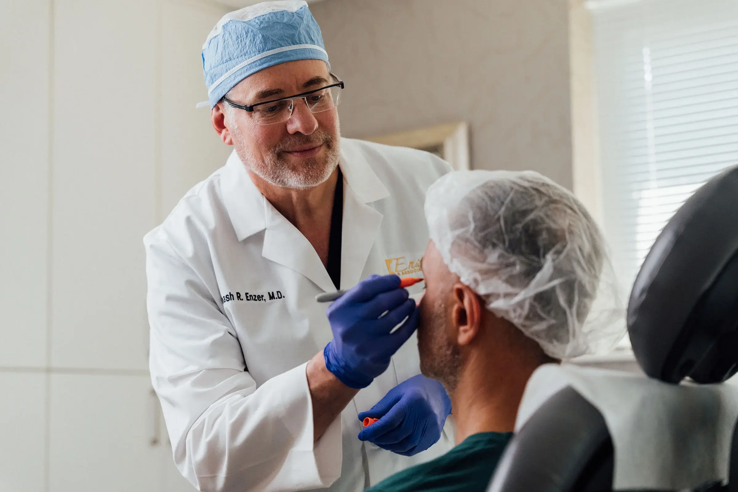 Dr. Enzer, wearing a white lab coat and blue surgical gloves, is marking the face of a patient in a surgical cap with a red marker during a pre-operative consultation.