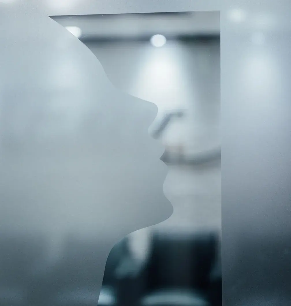 A close-up of a frosted glass panel featuring a cutout or etched silhouette of a face in profile, with a blurred, bright office interior visible through the glass.