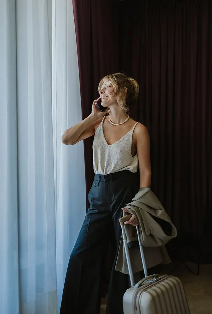 A smiling woman with blonde hair, wearing a pearl necklace, a light-colored tank top, and black pants, stands near a window talking on a cell phone while holding the handle of a suitcase and a beige jacket.