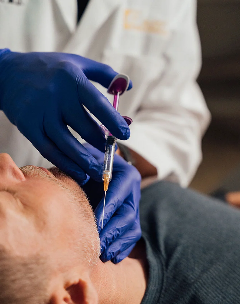 A medical professional wearing blue gloves and a white lab coat is administering an injection with a syringe to a male patient's neck area. - Kybella in Providence, RI