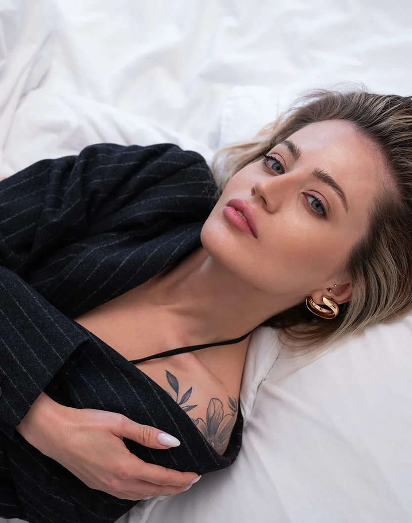 A portrait of a woman with blonde hair lying on white sheets, wearing a black pinstripe blazer and a black top, with a floral tattoo visible on her shoulder. - Lasers for Brownspot Tattoos in Providence, RI