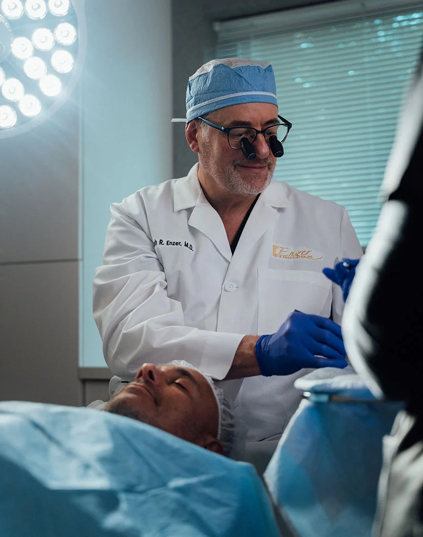 Dr. Enzer, wearing a surgical cap, loupes, and a lab coat, smiles while leaning over a male patient who is lying down covered by surgical drapes under a bright operating light. - Mole Skin Growth Removal in Providence, RI