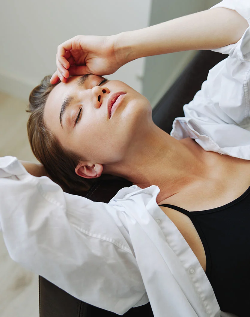 A woman with short brown hair, wearing a white unbuttoned shirt over a black top, is lying down with her eyes closed, gently resting her hand on her forehead. - Neck Liposuction in Providence, RI