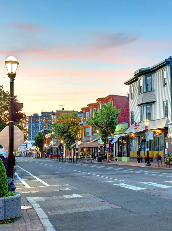 A wide street lined with historic row houses converted into shops and restaurants, illuminated by streetlights under a colorful sunset sky. - Out of Town Recovery in Providence, RI