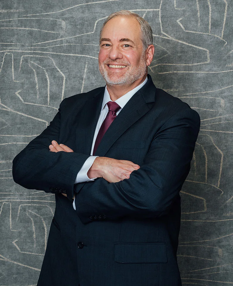 A professional headshot of Dr. Enzer, a man with gray hair and a beard, smiling with his arms crossed while wearing a dark suit and a maroon tie. He is standing against a gray wall with a subtle abstract white pattern.