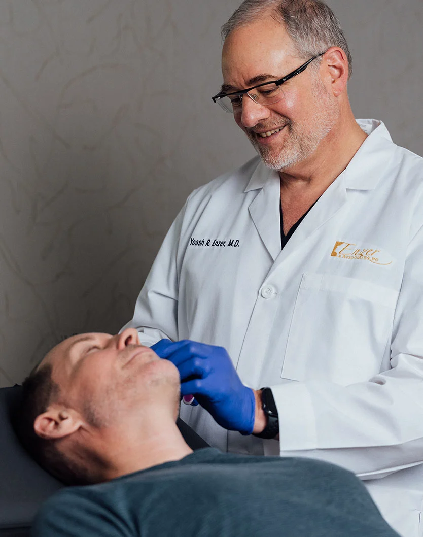 Dr. Enzer, wearing a white lab coat and glasses, smiles while examining the neck and jawline of a male patient who is lying down in a treatment chair. - Thread Lifts in Providence, RI