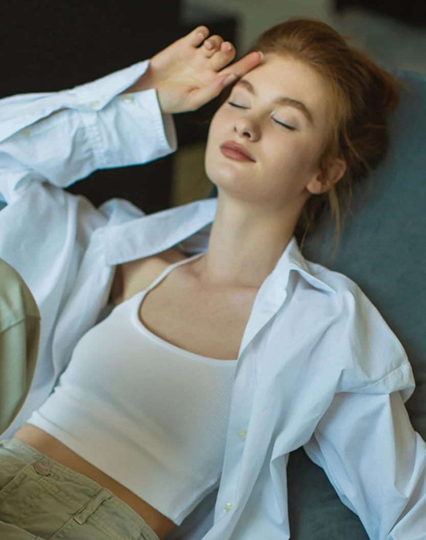 A woman with red hair, wearing a white tank top and an unbuttoned white shirt, is lying down with her eyes closed and her hand resting on her forehead. - Xanthelasma in Providence, RI